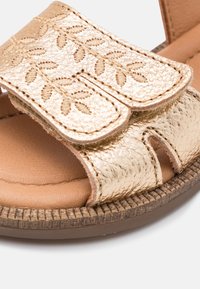 Złote metaliczne sandały z teksturowanym wzorem liścia na górnym pasku, z wyściełaną brązową wkładką i żłobioną podeszwą.