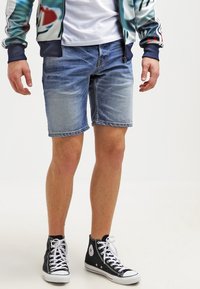Bermudas de mezclilla en azul claro con desvanecidos, combinadas con zapatillas de caña alta negras. Modelo vistiendo una camiseta blanca y una chaqueta con estampado.