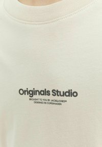 Ljusbeige bomullströja med rund halsringning. Har en svarttryckt logotext "Originals Studio" i fet stil. Slät textur.