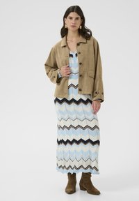 Jachetă din suede bej cu nasturi negri, asociată cu o rochie maxi cu model în zigzag în nuanțe de albastru, maro și crem. Botine maro până la gleznă.