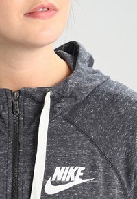 Šedá mikina na zip vyrobená z texturované látky, s bílým zipem, šňůrkami a výrazným bílým logem Nike na hrudi.