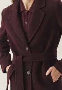 Burgundská bunda s měkkou texturou, širokými klopami, zapínáním na knoflíky, páskem v pase a slim fit střihem. Detaily knoflíků jsou v kontrastní barvě.