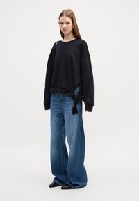 Felpa nera corta con dettaglio in cravatta sul lato, abbinata a jeans blu a vestibilità morbida. Il modello si trova davanti a uno sfondo semplice.