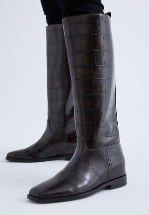 Botas altas hasta la rodilla de color marrón oscuro con textura de cocodrilo, usadas sobre pantalones negros sobre un fondo claro.