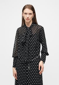 Γυναίκα που φοράει μαύρο διάφανο μπλουζάκι και φούστα με λευκές πουά, με δεμένο φιόγκο στο γιακά, ασημένια σκουλαρίκια κρίκους και ασημένια ζώνη με αλυσίδα.