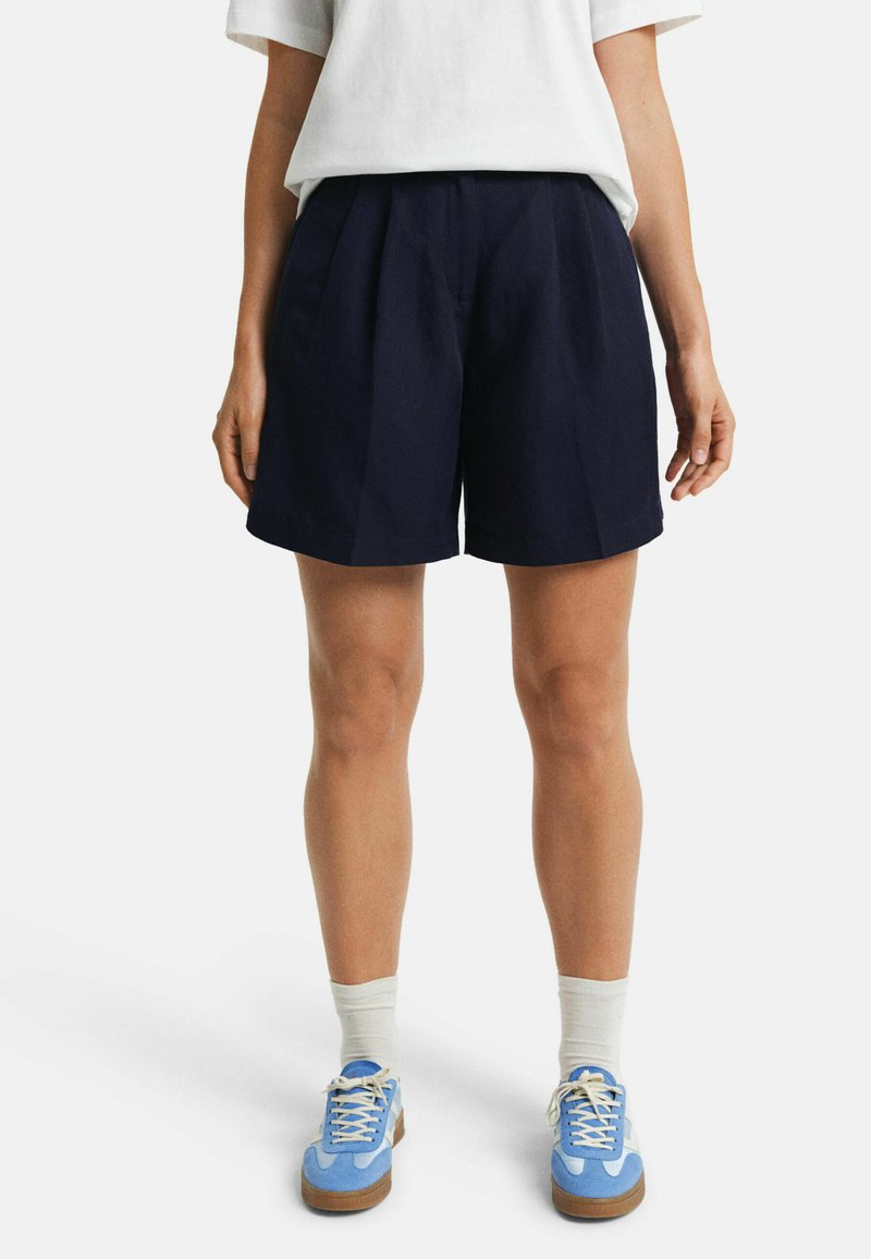 Shorts bleu marine, jusqu'aux genoux, avec des plis, fabriqués en tissu léger. Associés à un t-shirt blanc et des baskets bleues.
