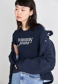 Námořnická modrá bavlněná tričko s nápisem "tommy jeans" vytištěným bílou barvou. Spárované s modrou prošívanou bundou a světlými modrými denimovými džíny.