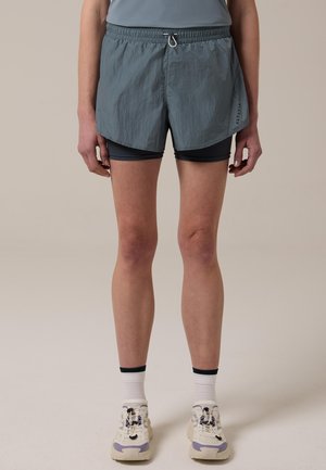 Donna che indossa pantaloncini da corsa grigi a strati, calzini bianchi e scarpe da ginnastica beige con dettagli neri e viola, in piedi davanti a uno sfondo neutro.