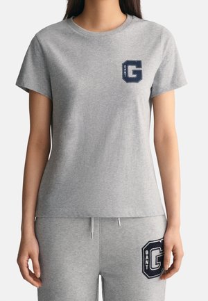 Kobieta ubrana w jasnoszary t-shirt z krótkim rękawem i pasujące spodnie dresowe, oba z granatowymi naszywkami z literami "GANT" na piersiach i udzie.