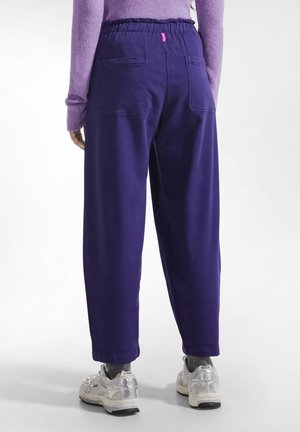 Person iført løse lilla bukser med baglommer, grå sokker, sølv sneakers og en lilla langærmet top, stående på en hvid gulv.