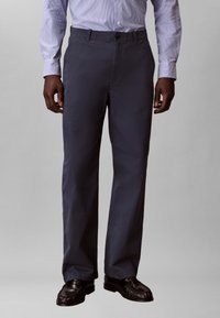 Άνδρας που φοράει σκούρο μπλε παντελόνι, μαύρα δερμάτινα μοκασίνια και ανοιχτό μπλε πουκάμισο με λωρίδες και μακριά μανίκια, στέκεται μπροστά σε απλό φόντο.