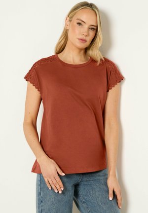 Vaaleahiuksinen nainen, joka pukeutunut ruosteenväriseen lyhythihaiseen paitaan, jossa on aaltoilevat pitsireunat, ja sinisiin farkkuihin, seisoo valkoista seinää vasten.