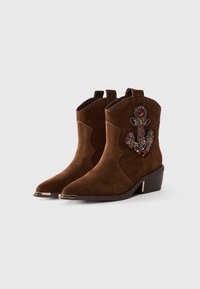 Brown suede ankle boots with chunky heels, metal toe caps, and decorative multicolored gemstone anchor on outer side.