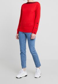 Czerwony sweter z dekoltem w łódkę, długie rękawy ozdobione czarnymi guzikami, zestawiony z jasnoniebieskimi dżinsami i białymi sneakersami.