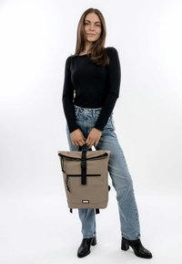 Zaino roll-top beige con hardware nero, tasca esterna, spallacci regolabili e materiale resistente. Il modello indossa jeans blu e una maglietta nera.