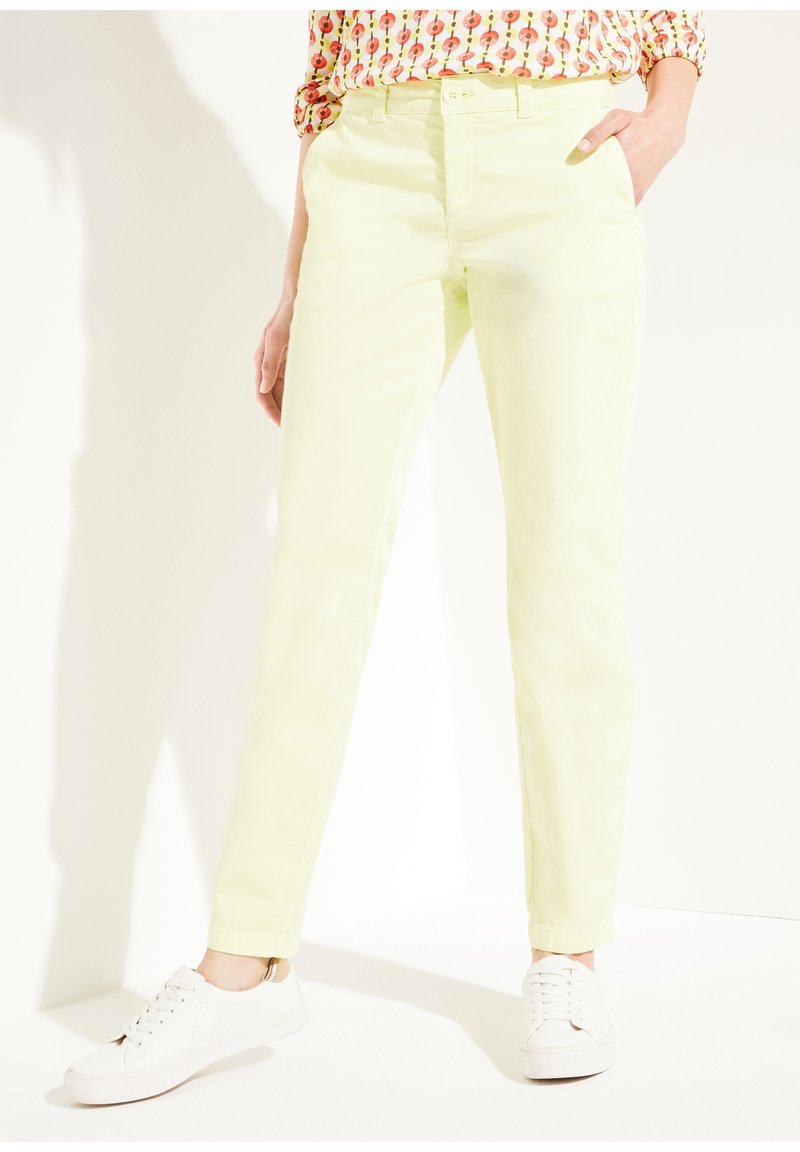 La personne porte un pantalon slim jaune pâle, des baskets blanches et une chemise à motifs avec une main dans la poche sur fond blanc.