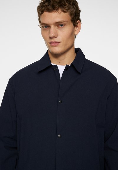 Marineblauwe blousejack met een puntige kraag, textuurstof en zwarte knopen. Ontworpen met een losse pasvorm en minimale details.