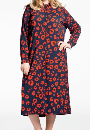 Žena v tmavě modrých midi šatech s dlouhým rukávem a výrazným červeným leopardím vzorem, stojící s jednou rukou na pasu a druhou volně u těla.