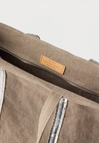 Μπεζ τσάντα καμβά με κλείσιμο με φερμουάρ, διακοσμημένη με ασημένιες παγιέτες στα πλάγια και δερμάτινη ετικέτα που αναγράφει τη μάρκα.