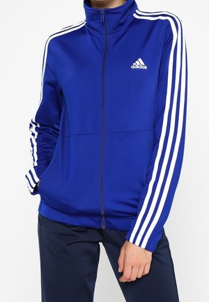 Personne portant une veste Adidas zippée bleue avec des rayures blanches sur les manches et le logo Adidas sur la poitrine, associée à un pantalon foncé.