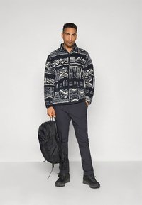 Flis jarek z abstraktnim mornarsko modrim in kremnim vzorcem, zasnova s polovičnim zadrgo, kombiniran s črnimi hlačami in črnim nahrbtnikom. Črna obutev.