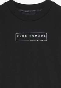 Czarny sweter o żebrowanej teksturze, z białym prostokątnym logo z napisem "CLUB NOMADE" i "SCOTCH & SODA" poniżej.