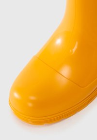 Botte en caoutchouc jaune avec une finition brillante. Tête de chaussure arrondie, couture surélevée au milieu, semelle texturée pour l'adhérence.