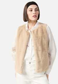 Beige fuskpälsväst med rundad halsringning och utan knäppningar, bärs över en vit skjorta med knappar. Det texturerade tyget visar en subtil lyster.