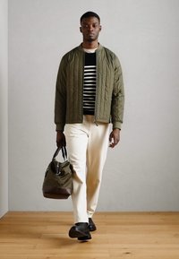 Giacca bomber trapuntata verde oliva su una maglietta a righe bianco e nera, abbinata a pantaloni crema e una borsa verde con dettagli marroni. Scarpe nere.