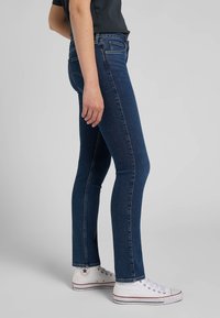 Skinny jeans in dark blue denim, featuring a high waist, slight stretch, and classic stitching. Paired with white high-top sneakers.