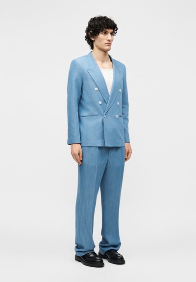Homme aux cheveux bouclés et foncés portant un costume croisé bleu clair, une chemise blanche et des chaussures noires, debout devant un fond uni.