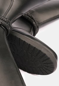 Bota de cuero negro con suela texturizada y cuentas decorativas negras en el tobillo. Presenta un acabado suave y costuras reforzadas.