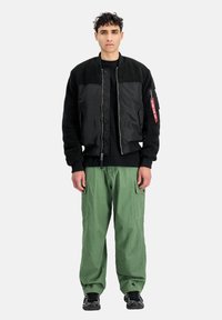 Mladý muž stojící čelem, oblečený v černé bombérce, černém tričku, zelených cargo kalhotách a černých teniskách na bílém pozadí.