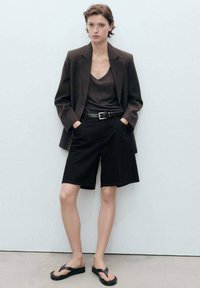 Blazer marron foncé sur un débardeur sombre, associé à un short noir et des tongs noires ; tissu lisse et design minimaliste.