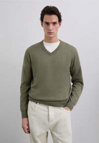 Suéter verde con cuello en v hecho de tela suave, con mangas largas y puños acanalados, combinado con pantalones blancos. Sin patrones o detalles destacables.