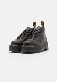 Botas de tobillo de cuero negro con suela gruesa y estriada, costuras amarillas y cordones negros. Presentan una lengüeta con la marca en el talón.