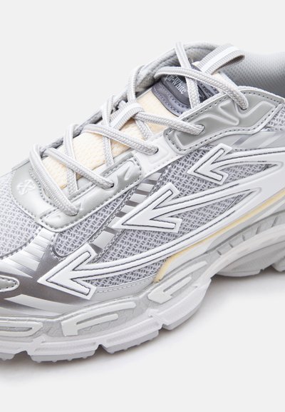 Κοντινή λήψη γκρι και λευκού δίχτυνου αθλητικού παπουτσιού με επικαλύψεις σε σχήμα βέλους και παχύ, μαλακό πέλμα, δεμένο με γκρι κορδόνια.