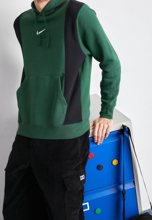 Osoba ubrana w zielono-czarną bluzę Nike z kapturem i czarne spodnie opiera się o niebieską szafkę z kolorowymi okrągłymi magnesami.