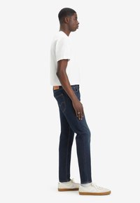 Jeans ajustados de mezclilla azul oscuro con un diseño clásico de cinco bolsillos, combinados con una camiseta blanca de manga corta y zapatillas blancas.