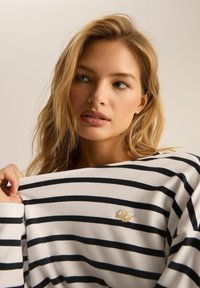 Pasykowa bluzka z długim rękawem w kolorze białym i granatowym, z miękką fakturą i haftowanym logo w kolorze złotym. Luźny krój z szerokim dekoltem.