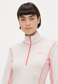 Ροζ ανοιχτό ροζ τοπ με φερμουάρ στο ένα τέταρτο, με λουλουδάτο σχέδιο και αντίθετες σκουρότερες ροζ λεπτομέρειες. Διαθέτει ψηλό γιακά και λογότυπο στο στήθος.