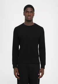 Black ribbed long-sleeve shirt with a crew neckline and cuffed sleeves, worn with matching black pants, displayed on a model.