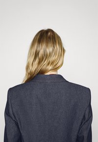 Personne aux longs cheveux blonds porte un blazer gris foncé, tournée de dos sur un fond clair uni.