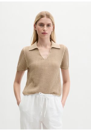 Blondynka ubrana w beżowy krótki sweter z kołnierzykiem i białe spodnie ze sznurkiem, stoi z rękami w kieszeniach.