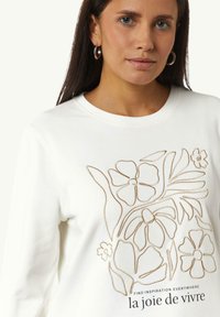 Witte sweatshirt met een goud geborduurd bloemenontwerp en tekst aan de onderkant. Ronde halslijn en een gladde textuur.