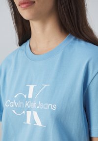 Gaiši zils kokvilnas T-krekls ar baltu logo, kurā ir teksts "Calvin Klein Jeans", ar apaļu kakla izgriezumu, īsām piedurknēm un mīkstu tekstūru.