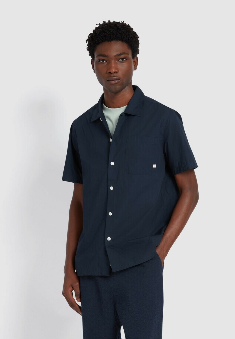 Camicia navy a maniche corte con colletto alla coreana, taschino sul petto e bottoni bianchi. Tessuto liscio, indossata sopra una maglietta azzurra chiara.