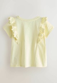 Halvány sárga kisgyermek póló rövid ujjakkal és fodros szegéllyel, amely a vállaktól a póló elülső oldalának oldalai mentén fut le.