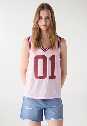 Giovane donna con lunghi capelli biondi che indossa una canotta sportiva rosa senza maniche con il numero "01" e pantaloncini di jeans strappati, su uno sfondo semplice.