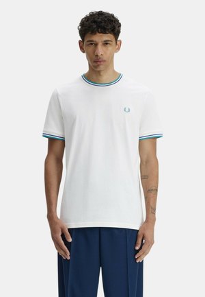 Giovane uomo con indosso una maglietta bianca con bordo a strisce blu e verde e pantaloni blu, in piedi davanti a uno sfondo chiaro uniforme.